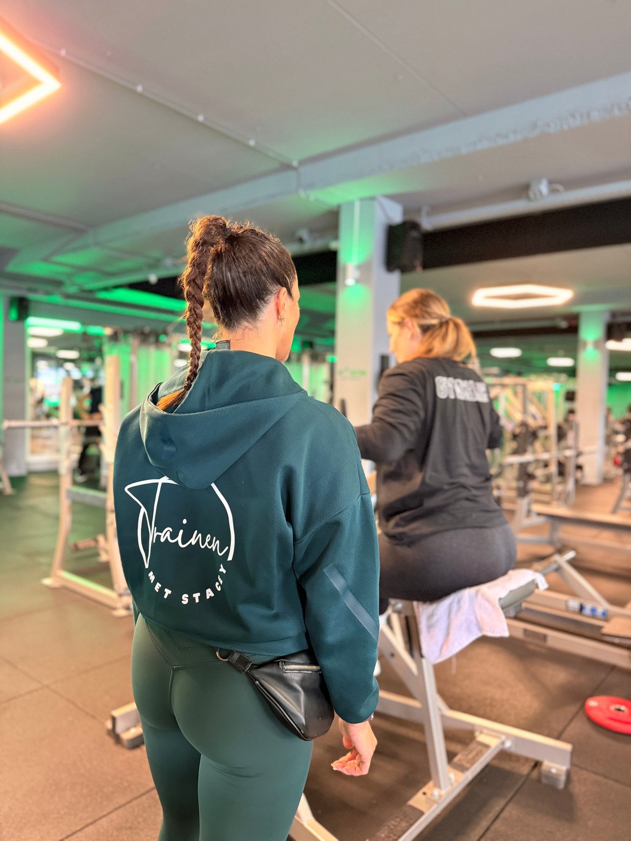 Stacey in haar Trainen met Stacey-hoodie begeleidt een klant in de sportschool